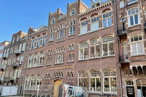 Historisch schoolgebouw van baksteen met sierlijke details en grote boogramen, fietsen geparkeerd voor de ingang.
