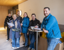 Vijf personen poseren samen in een lichte, moderne binnenruimte met tafels, stoelen en een grote vaas met kleurrijke bloemen. Eén persoon zit op een hoge kruk; de anderen staan eromheen.