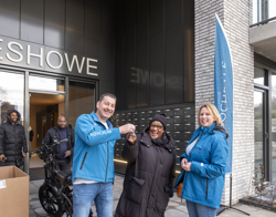 Twee personen staan voor de entree van het gebouw Eshowe, waarbij een sleuteloverdracht plaatsvindt. Naast hen staat een grote blauwe Rochdale-vlag en op de achtergrond is een banner te zien met een welkomsboodschap.