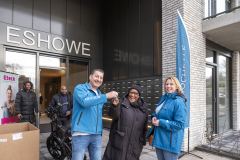 Twee personen staan voor de entree van het gebouw Eshowe, waarbij een sleuteloverdracht plaatsvindt. Naast hen staat een grote blauwe Rochdale-vlag en op de achtergrond is een banner te zien met een welkomsboodschap.